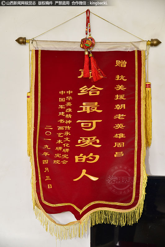      身后的锦旗，是2019年中华英雄传承文化研究会、中国军建书画艺术研究院的人前来看望时，献给老人的，经历过战争，才知道和平的可贵，也让老人更加珍惜现在的生活。